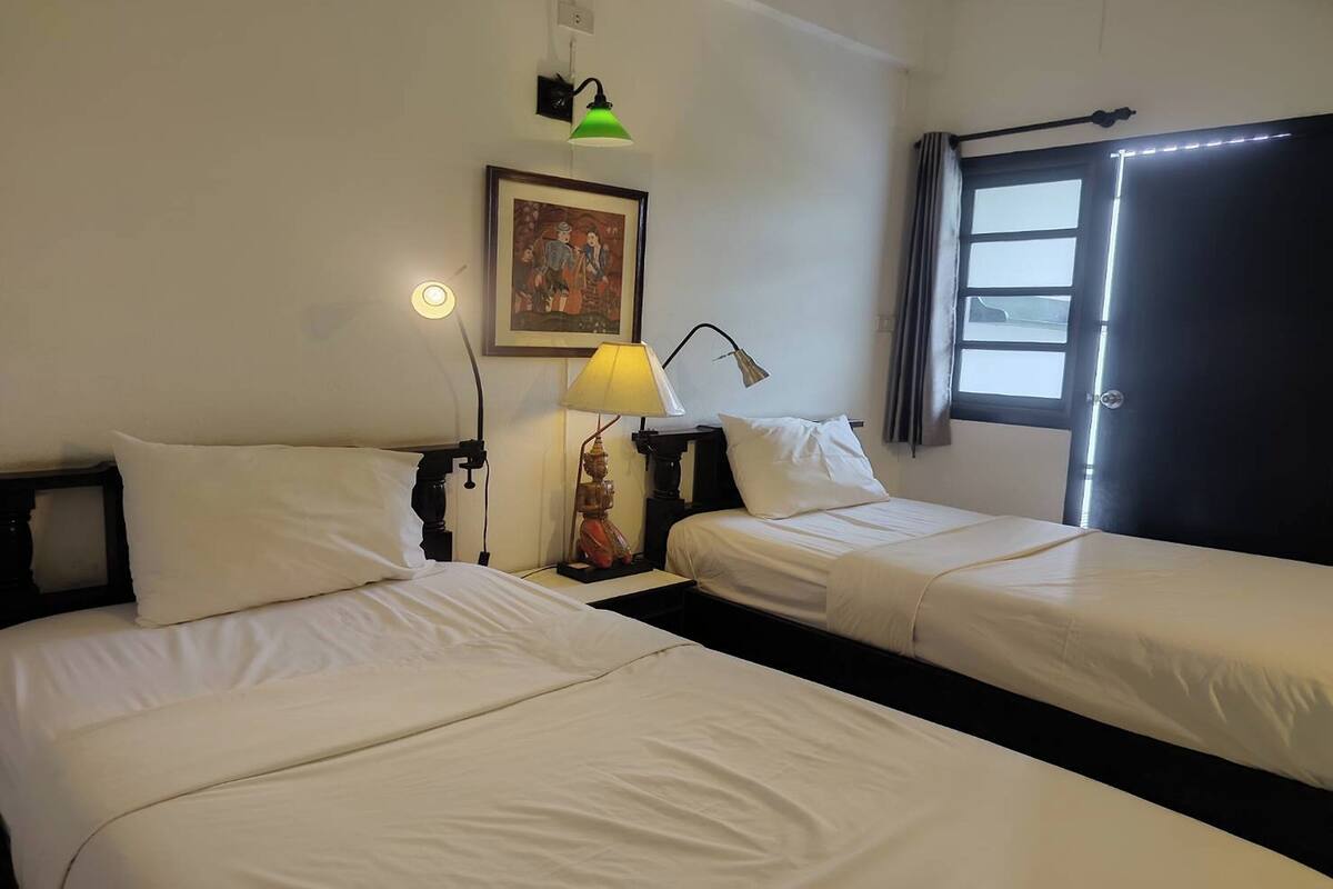 Habitación de hotel con dos camas individuales, una mesita de noche con lámpara, un cuadro en la pared y una ventana con cortinas.