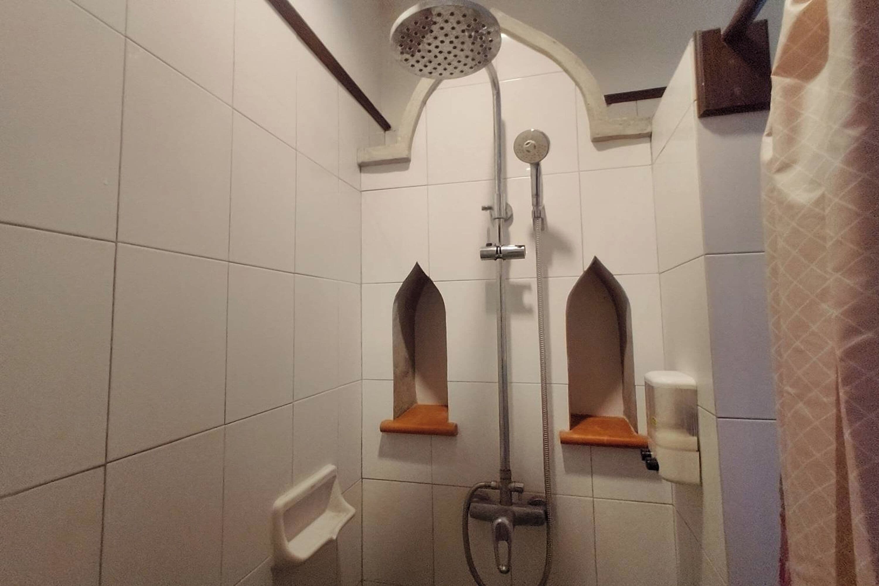 A shower with a handheld showerhead and a wall-mounted faucet. There are two small shelves with soap dispensers.