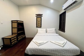 A neatly made bed with white linens and towels folded at the foot, a wooden storage unit with drawers, a wall-mounted air conditioning unit, and a window with a closed dark blind. - Orchid Hostel Near Bumrungrad Hospital and Nana Market (Bangkok)