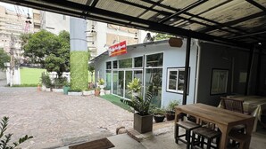Un espace extérieur aménagé, abrité, avec une table et des chaises en bois, des plantes en pot et un bâtiment doté de grandes fenêtres et d’une enseigne.