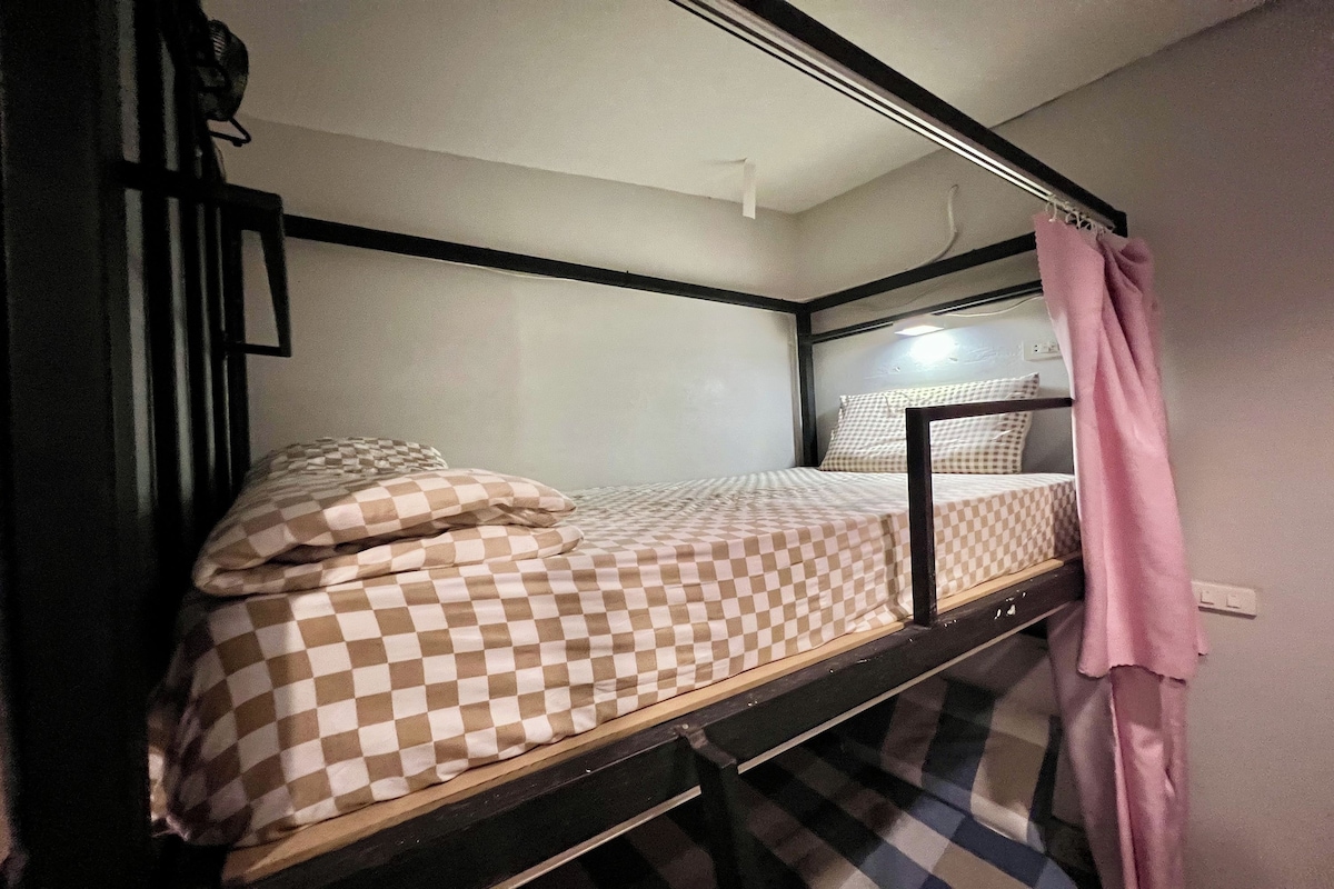 A bunk bed with a checkered bedspread and pillows, a black metal frame, and a pink curtain.