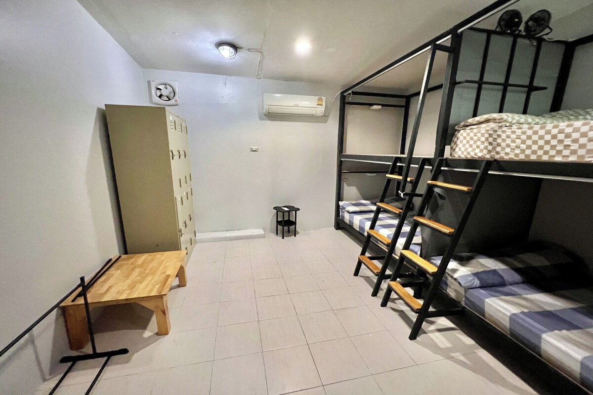 A dormitory room with bunk beds, a wooden desk, a small stool, and an air conditioning unit.