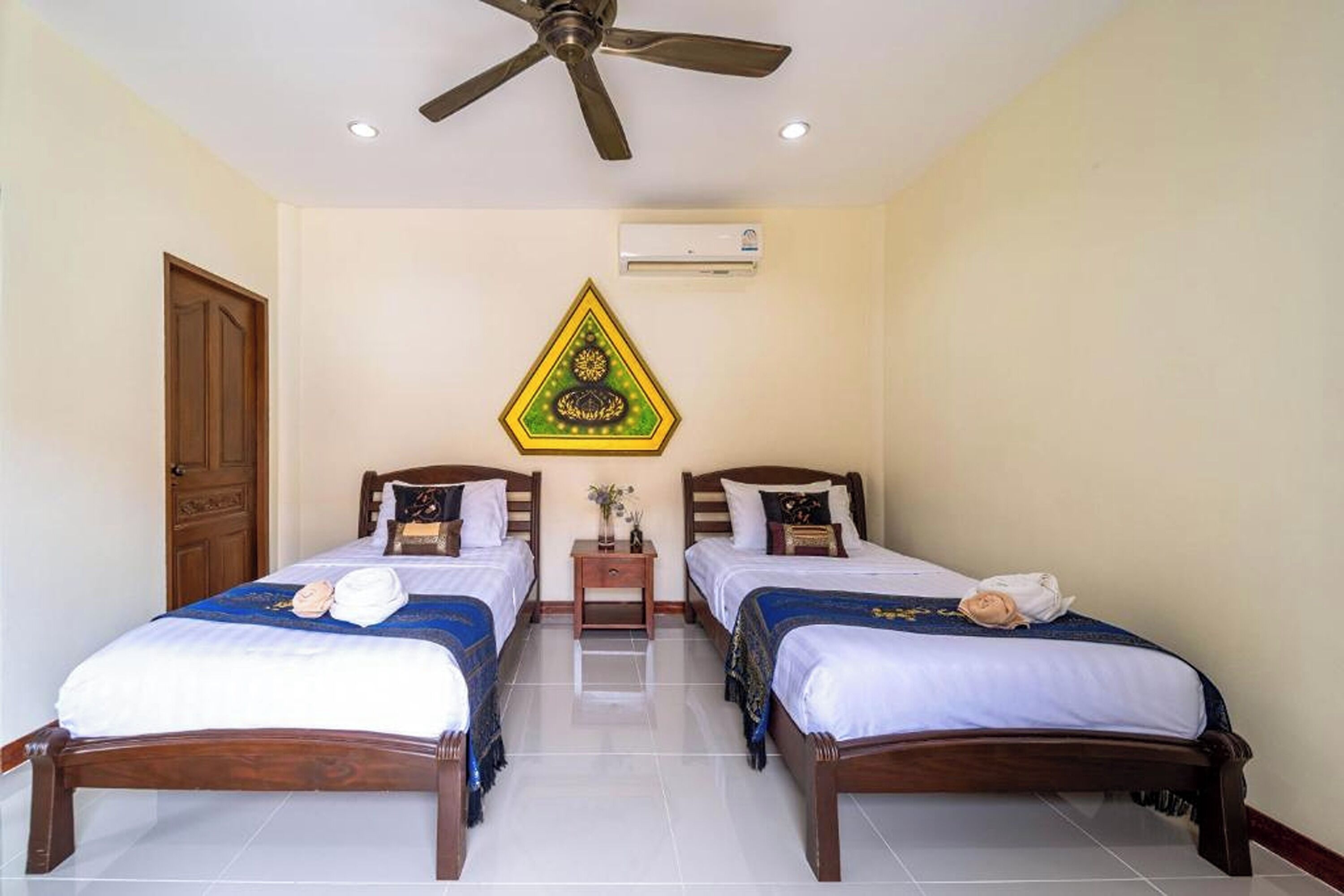 Habitación de hotel con dos camas de madera, un ventilador de techo, un equipo de aire acondicionado y un tapiz decorativo.