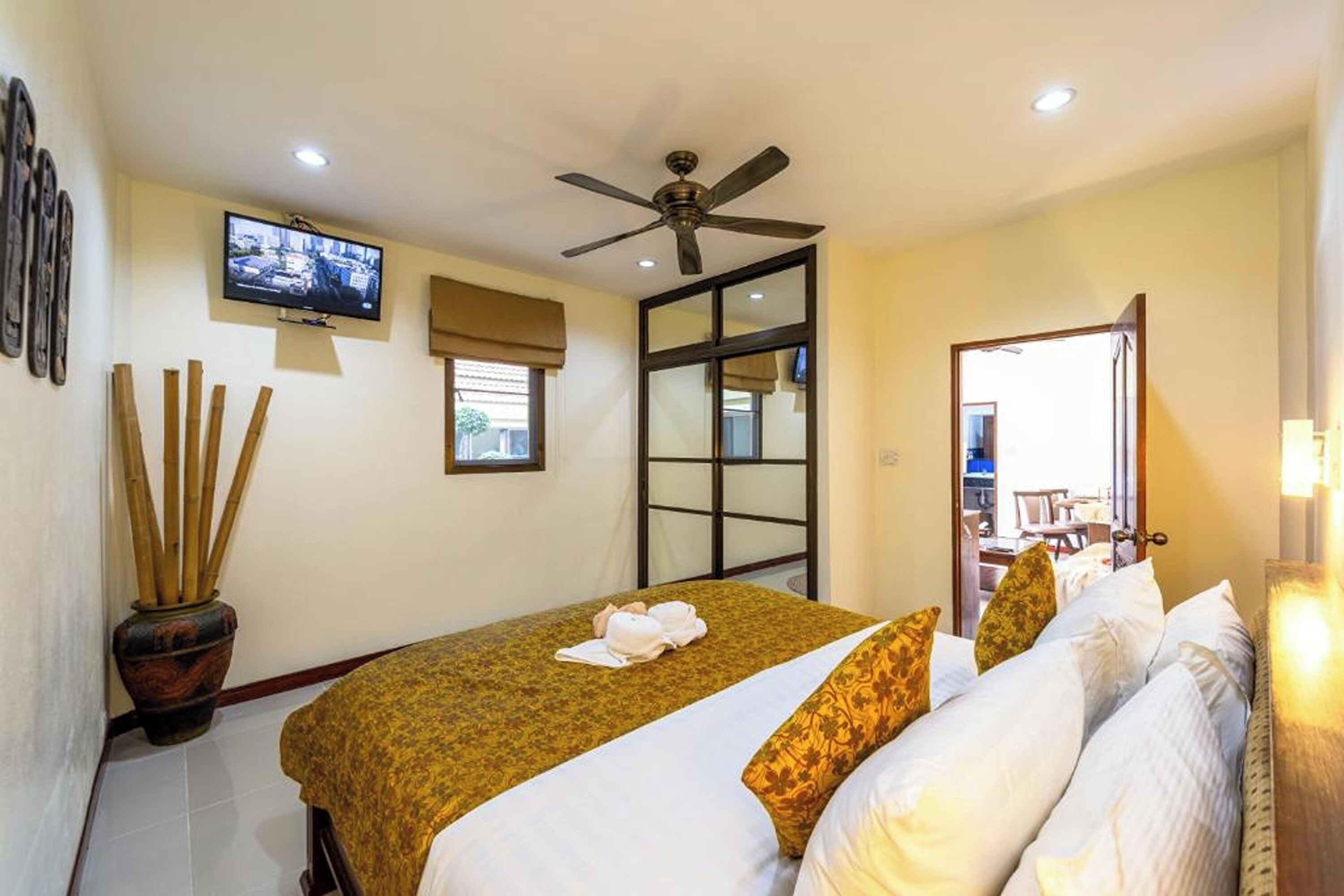 Habitación de hotel con cama, ventilador de techo, televisor montado en la pared, ventana con persianas, puerta y una planta en maceta.