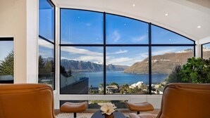 A room with a large panoramic window offering a view of a lake and mountains. There are two leather chairs and a small table with a vase.