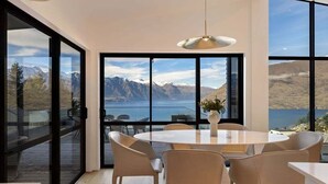 A modern dining area with a large round table, beige chairs, and a pendant light. It features expansive windows offering a view of mountains and a body of water.