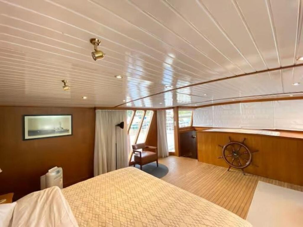 A cabin with a bed, a desk, a ship's wheel, and a window with curtains.