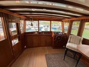 Een bootcabine met houten inrichting, een stuurwiel en uitzicht op het water.