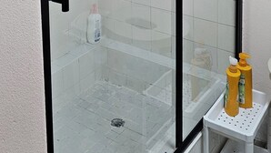 Ducha envuelta en vidrio, con cabezal de ducha de mano, pared de azulejos blancos y un pequeño estante con artículos de aseo.