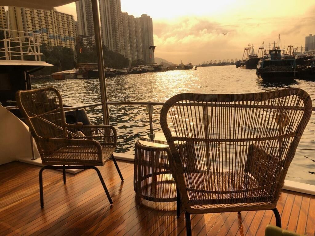 Ein Bootsdeck mit Korbmöbeln, einem kleinen runden Tisch und Blick auf eine Stadtlandschaft und Wasser.