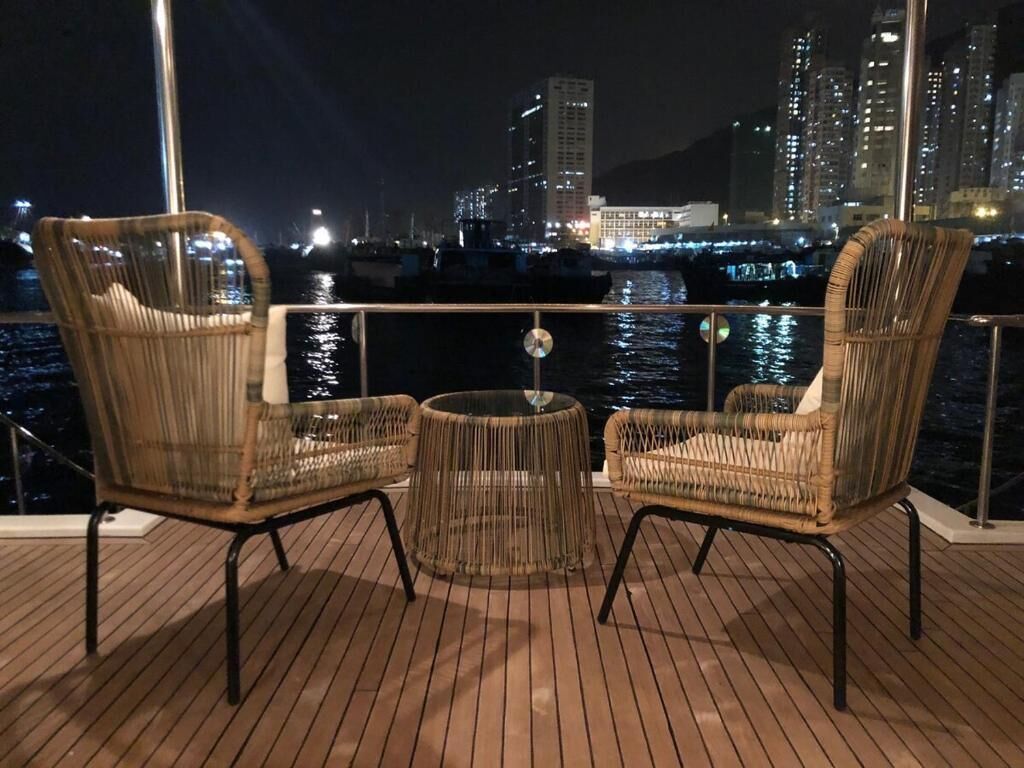 Een terras met rieten stoelen en een ronde tafel, met uitzicht op een jachthaven met boten en stadsgebouwen in de nacht.