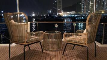 Een terras met rieten stoelen en een ronde tafel, met uitzicht op een jachthaven met boten en stadsgebouwen in de nacht.