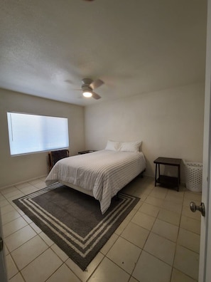 A bedroom with a bed, a ceiling fan, a window, a nightstand, a small table, and a laundry basket.