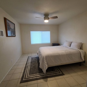 Un dormitorio con cama, ventilador de techo, ventana con persianas, alfombra y un cuadro en la pared.