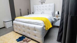 A bedroom with a tufted headboard, a yellow throw blanket, a white bedspread, and a small bedside table with a lamp. - Kilimani Homes (Nairobi)