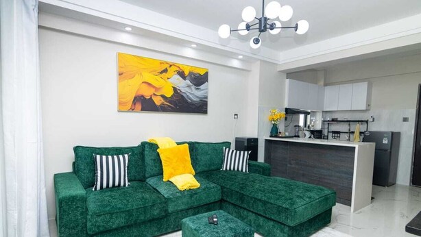 A modern living room with a green sectional sofa, a small ottoman, and a kitchen area in the background. - Kilimani Homes (Nairobi)