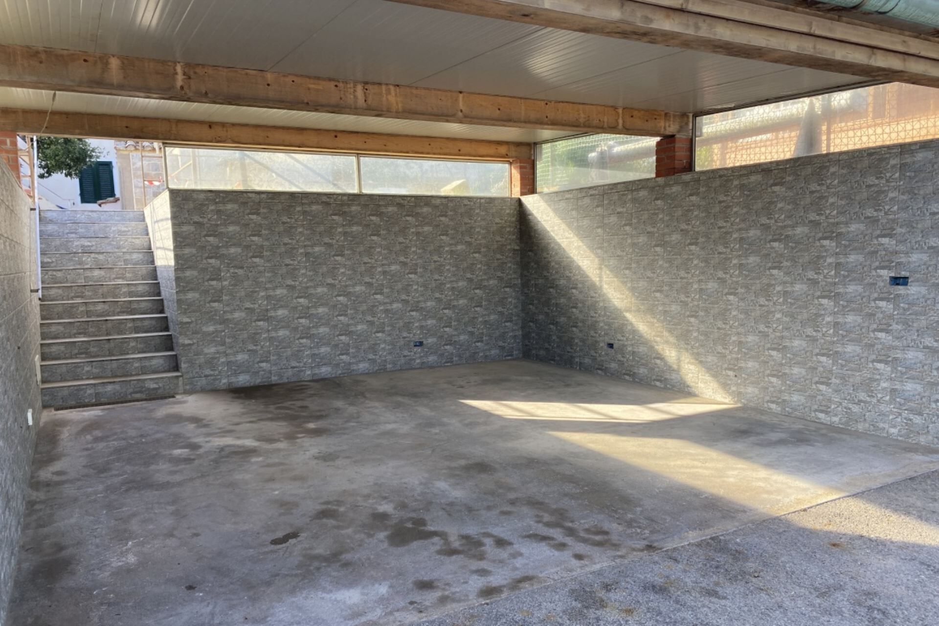 A room with a concrete floor, stone walls, and a wooden ceiling. There is a window on the left side, and a staircase leading to an upper level.