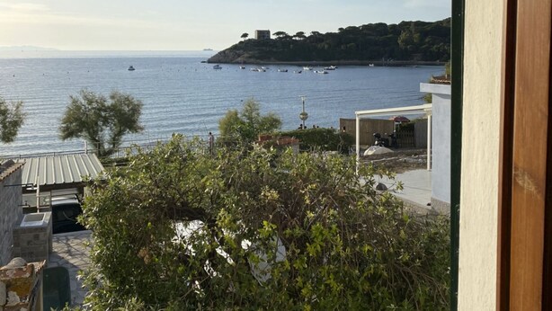 A coastal view with boats on the water, a hill with buildings, and a balcony with greenery.