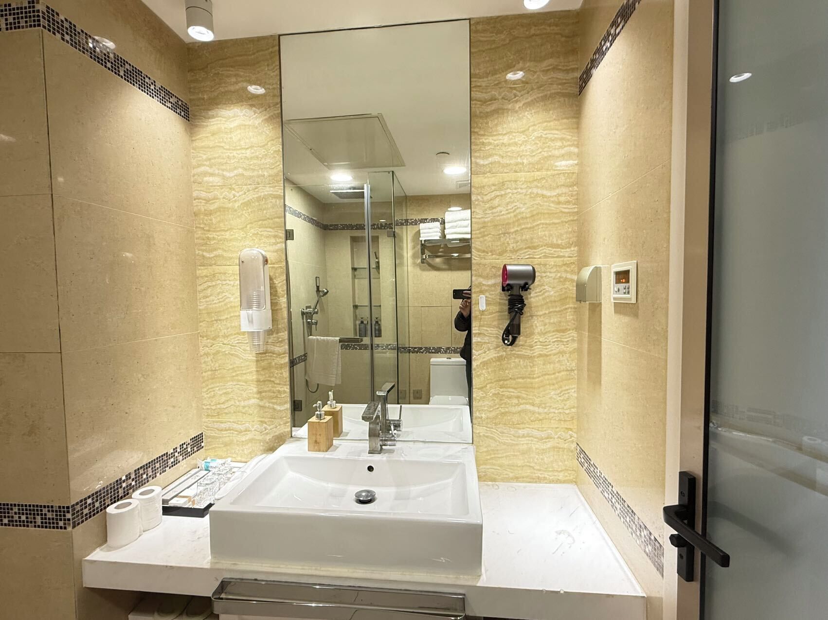 A modern bathroom with a large mirror, a sink, a handheld showerhead, and a hairdryer mounted on the wall.