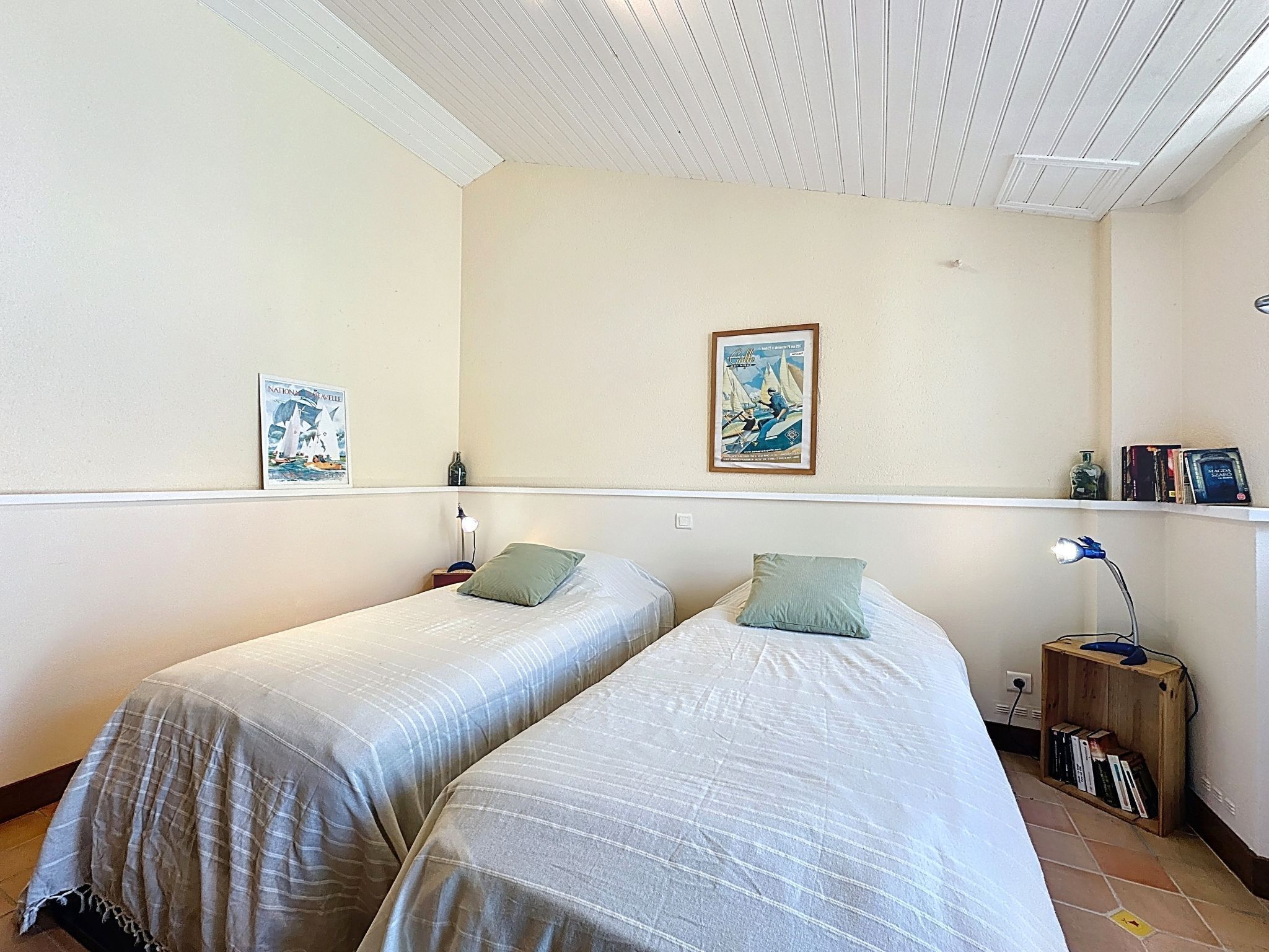 A room with two single beds, a small shelf with books, a framed picture, and a lamp on a nightstand.
