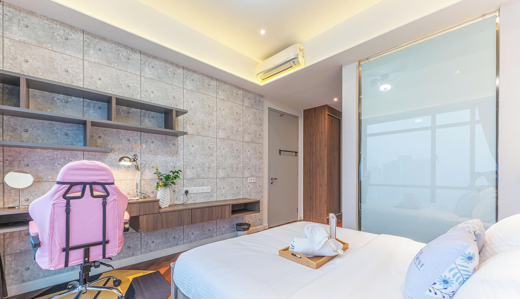 Ein modernes Hotelzimmer mit einem Bett, einem Schreibtisch mit pinkfarbenem Stuhl, einer kleinen Pflanze und einem großen Fenster.