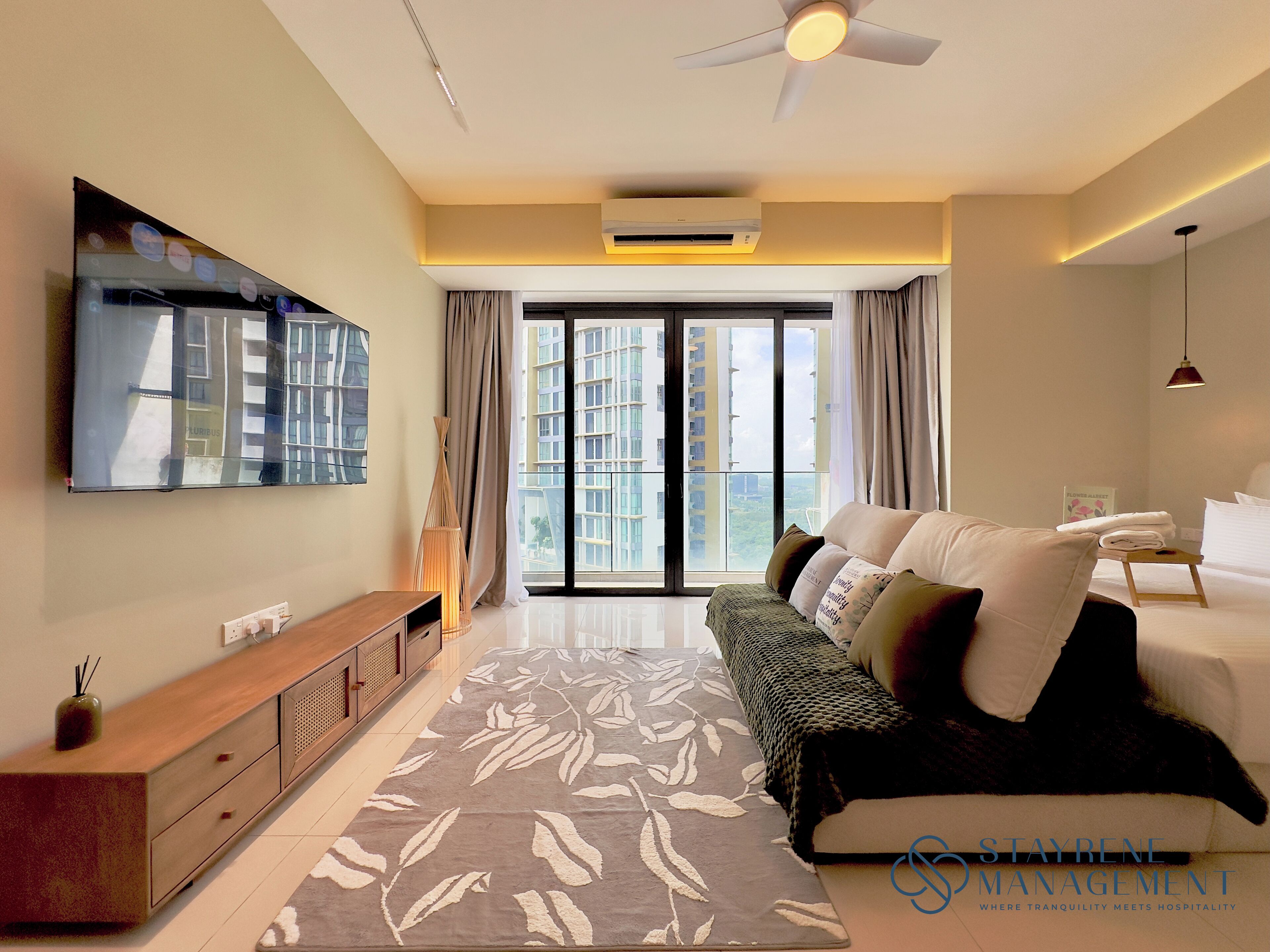 A modern hotel room with a large bed, a sofa, a TV mounted on the wall, and a view of the city through the windows.