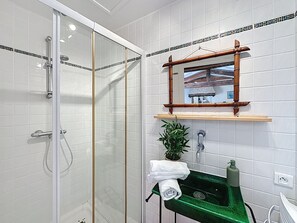 Ein Badezimmer mit einer gläsernen Duschkabine, einem grünen Waschbecken, einem Spiegel mit Holzrahmen und einer kleinen Pflanze.