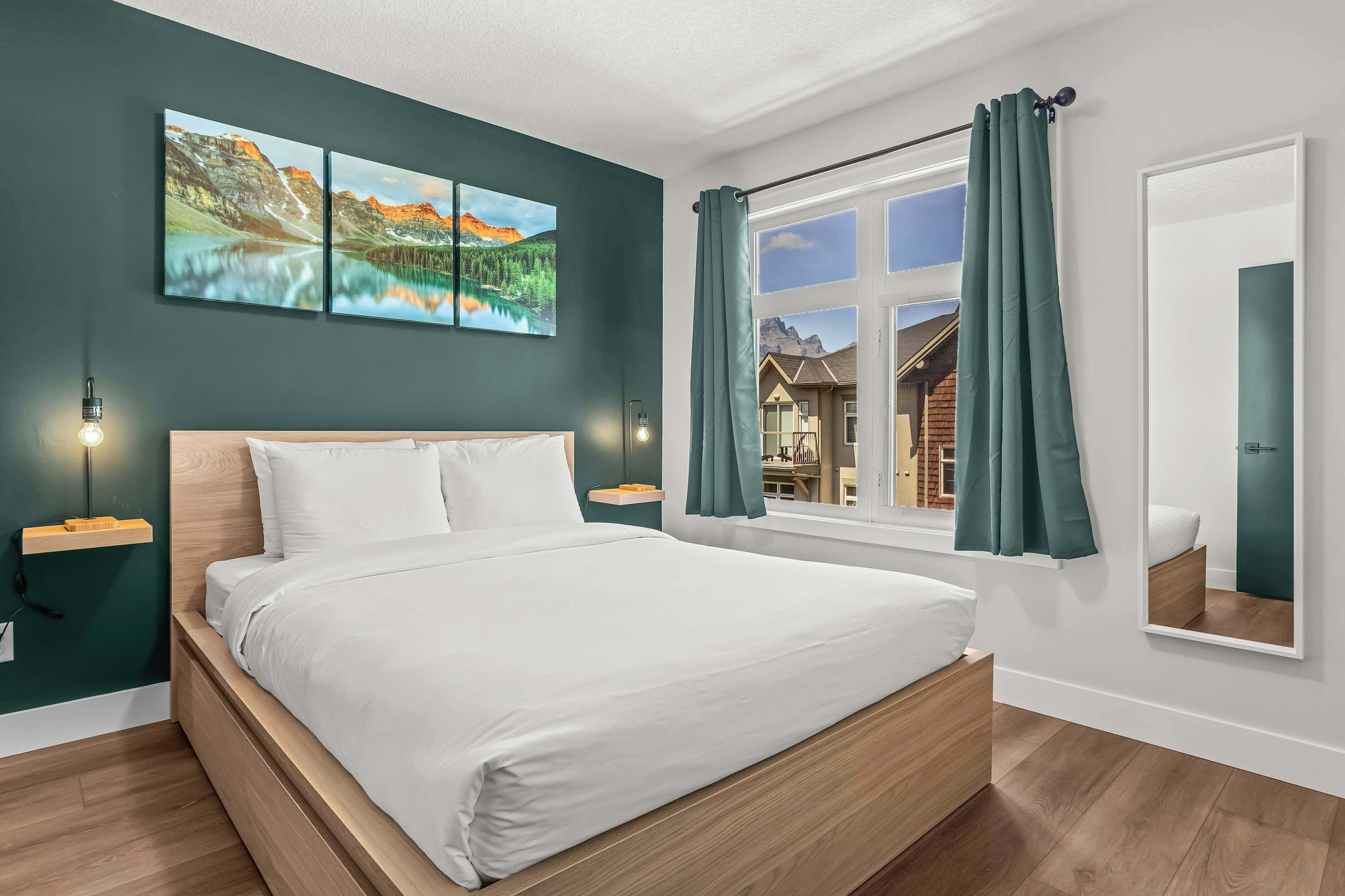 A modern bedroom with a wooden bed, a large window with curtains, and a view of a mountain house.