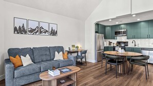 Een moderne woonkamer met een grijze bank, een houten salontafel, en een eethoek met een ronde tafel en stoelen. Op de achtergrond is een keuken met turquoise kastjes en roestvrijstalen apparatuur te zien.