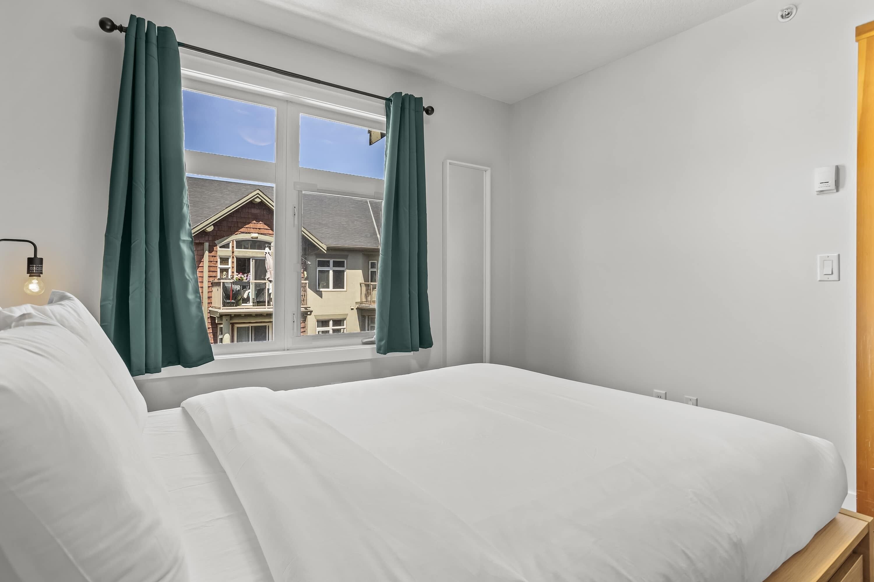 A bedroom with a large bed, a window with curtains, and a view of buildings outside.