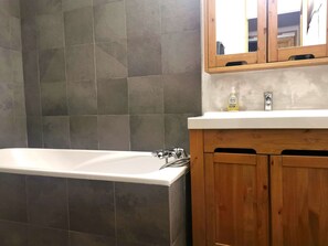 Un baño con bañera, baldosas grises, armarios de madera y un espejo sobre el lavamanos.