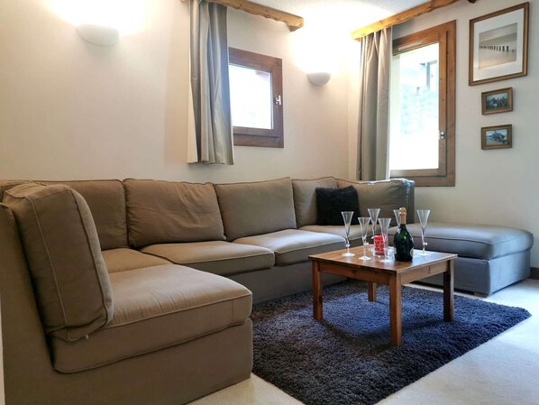 Una sala de estar con un sofá seccional color beige, una mesita con vasos y una botella, un tapete azul y una ventana con cortinas.
