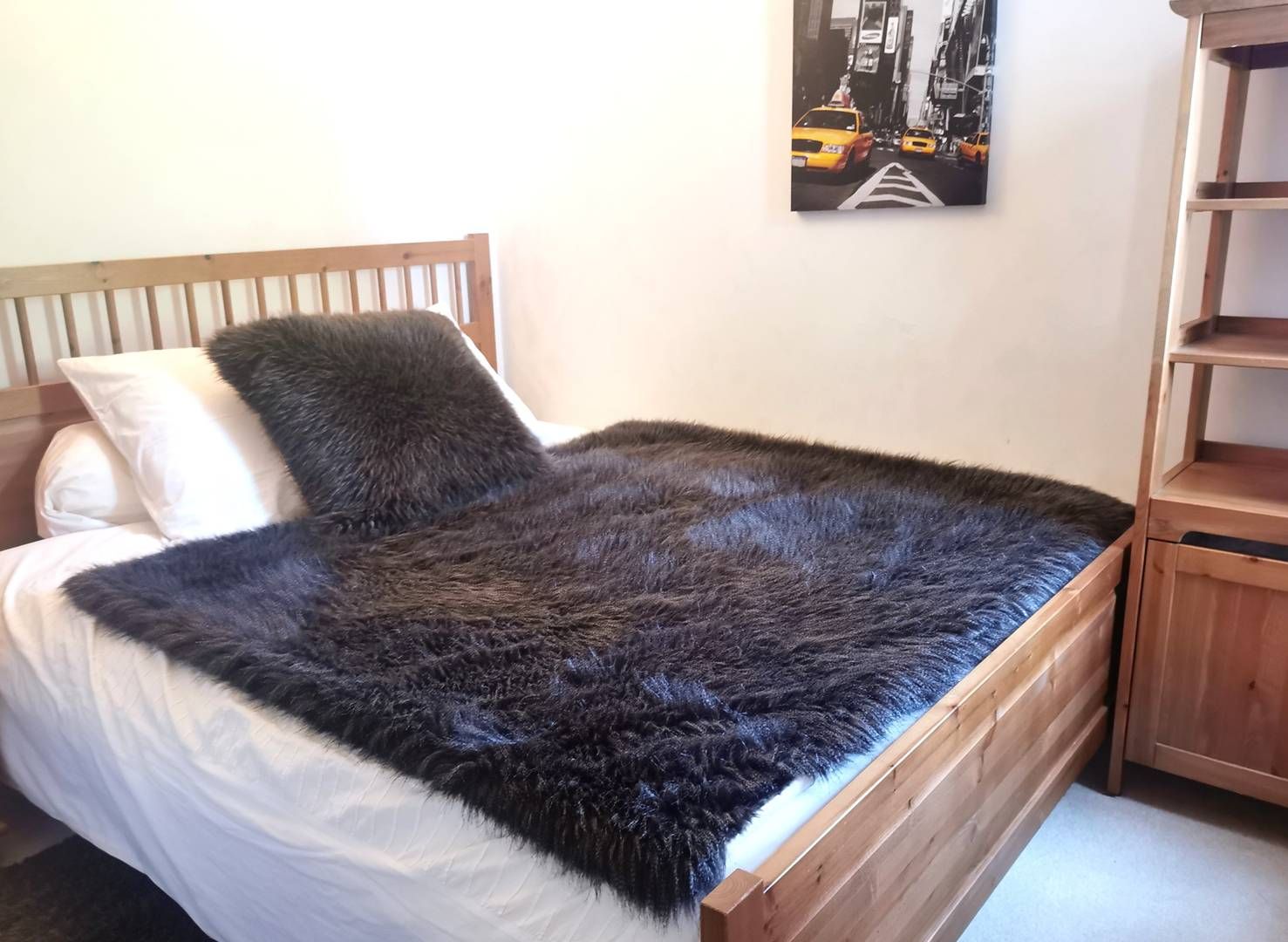 A bed with a white mattress and a dark, furry blanket. There is a wooden headboard and a white pillow. A painting of a city street is on the wall above the bed.