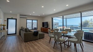 A spacious living room with a sofa, dining table, and chairs, large windows with a view of the sea, and a flat-screen TV on the wall.