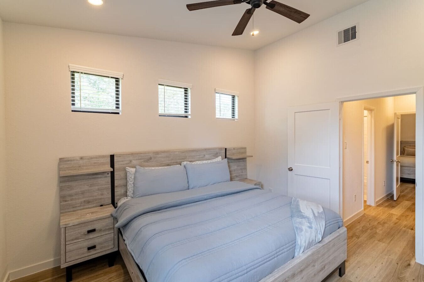Une chambre à coucher comprenant un lit, des tables de chevet, un ventilateur de plafond et une porte donnant sur une autre pièce.