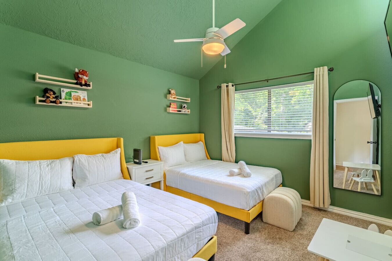A room with two beds, a ceiling fan, a window with curtains, a small table, and a mirror.