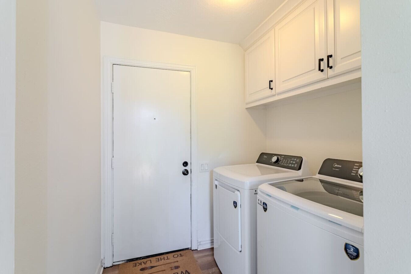 A laundry room with a white door, two stacked washing machines, and white cabinets above.