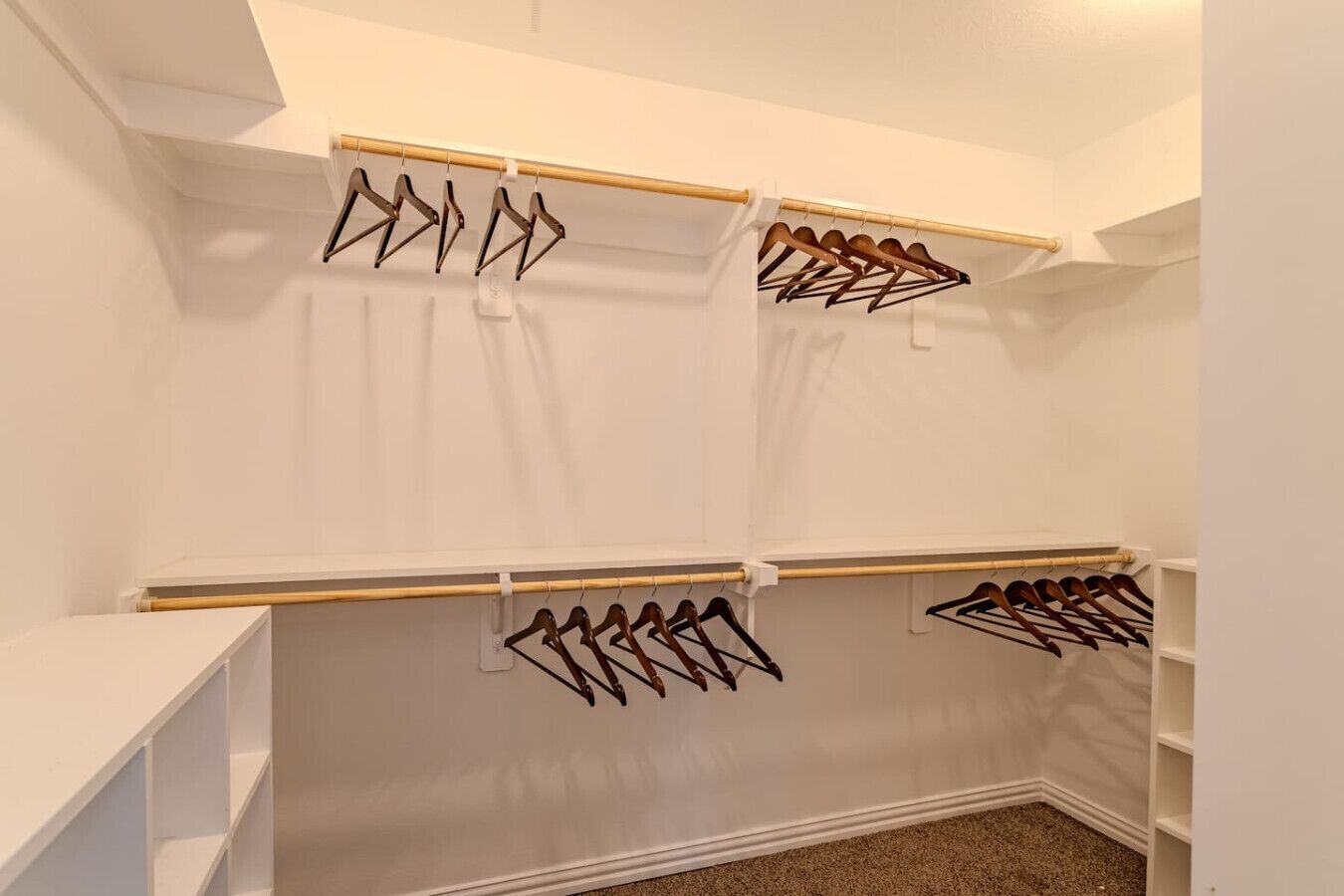 A walk-in closet with multiple wooden hangers on racks, white walls, and a carpeted floor.