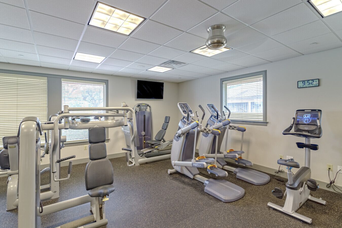 Un gimnasio equipado con diferentes máquinas de ejercicio, un reloj digital y ventanas con persianas.