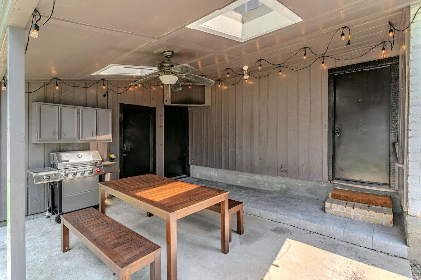 Un patio al aire libre con una mesa y bancos de madera, una parrilla y luces colgantes.