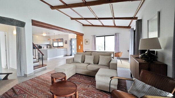 A spacious living area with a sofa, ottoman, and coffee table, wooden beams on the ceiling, and a view into the kitchen. - Villa Sun and Sea | Pool n' 2min walk to the Beach (Ponta Delgada)