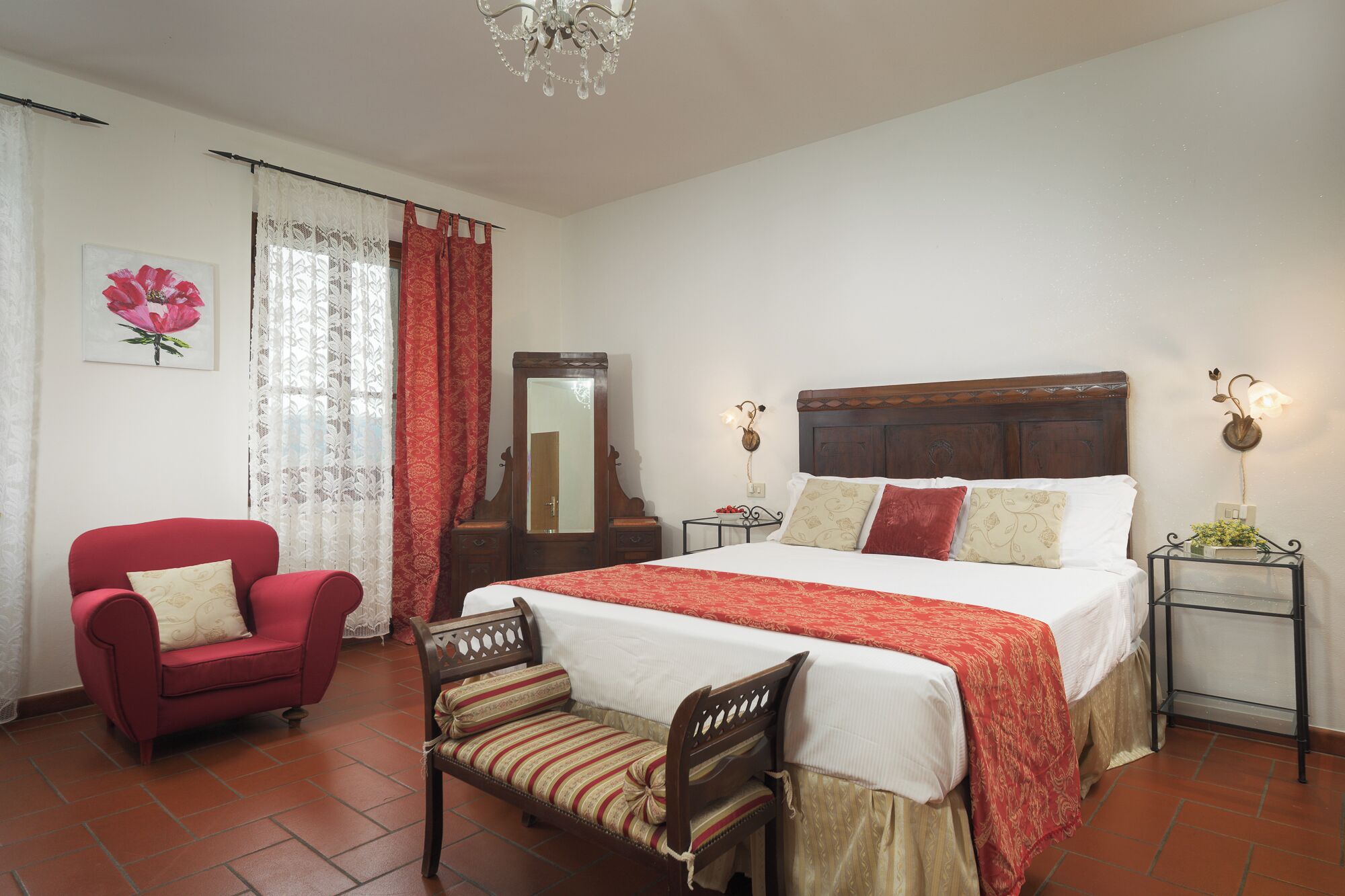 A bedroom with a large bed, a red armchair, a wooden chair, a wooden dresser, and a mirror.