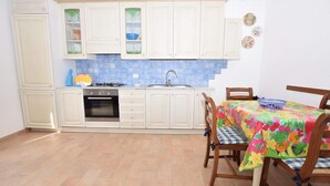Une cuisine avec des meubles blancs, une cuisinière à gaz, un four et un évier. On y trouve une table à manger avec des chaises et une nappe colorée.