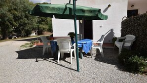 Un espace extérieur aménagé avec une table et des chaises sous un parasol vert, entouré d’un sol en gravier et d’un bâtiment blanc.