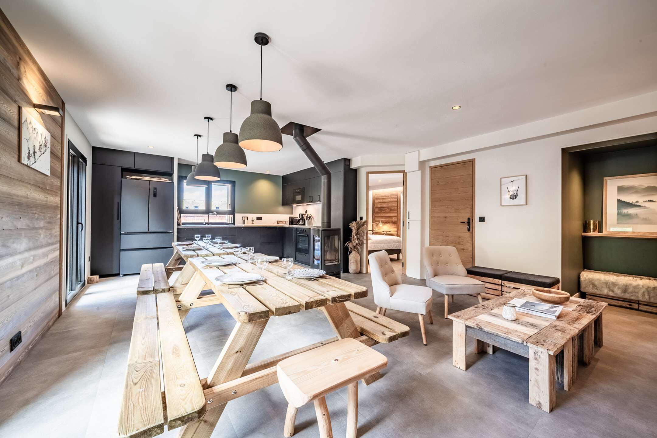 A modern kitchen with a long wooden table and benches, a dining area with a white chair, and pendant lights.