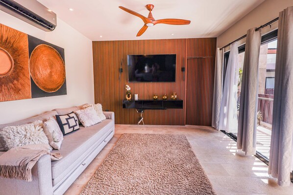 A modern living room with a flat-screen TV mounted on a wooden panel wall, a ceiling fan, a sofa with decorative pillows, and a large rug.