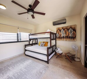 Ein Zimmer mit Etagenbett, Deckenventilator, Wandklimaanlage, Standventilator und einem Fenster mit Jalousien.