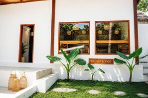 Un bâtiment blanc avec une enseigne indiquant « KELAPA LODGE », une porte en bois et deux grandes fenêtres ornées de guirlandes lumineuses.