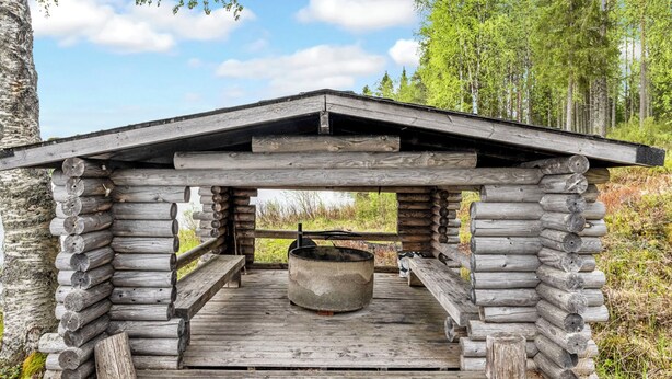 A rustic wooden cabin with a central stone fireplace, surrounded by a wooden deck and a natural forest backdrop. - Lumikenttien kutsu by Interhome (Kuusamo)