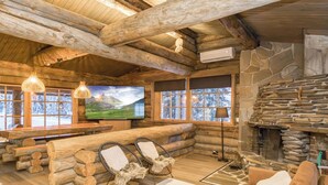 A log cabin interior with a stone fireplace, wooden ceiling beams, a large TV, and a cozy seating area. - Lumikenttien kutsu by Interhome (Kuusamo)
